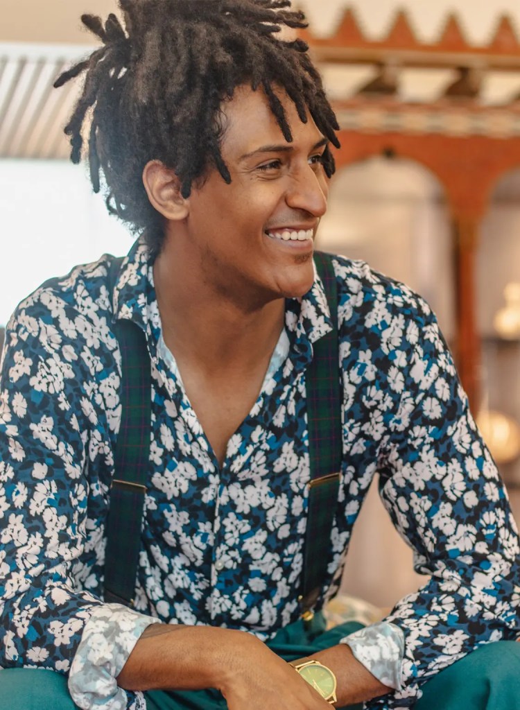 Model in blue shirt with white florals and suspenders, smiles and rests his forearms on his legs