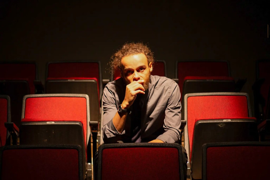 Man sits alone in theatre chairs with the spotlight on him as he is deep in thought
