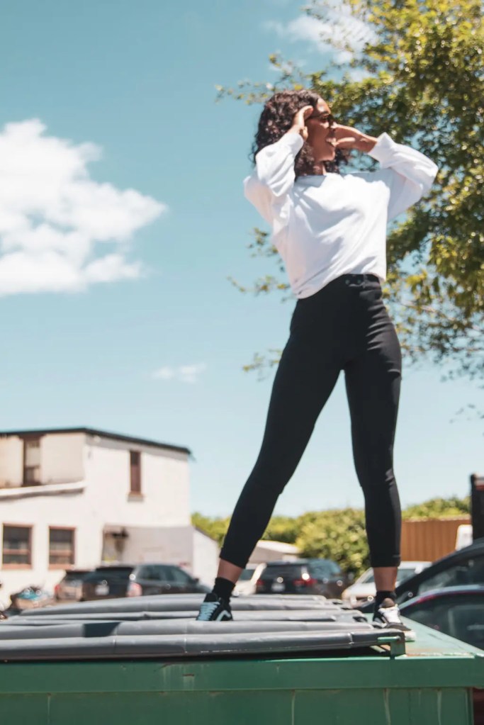 woman fixing her hair on top of a garbage bin wearing black pants and a white top