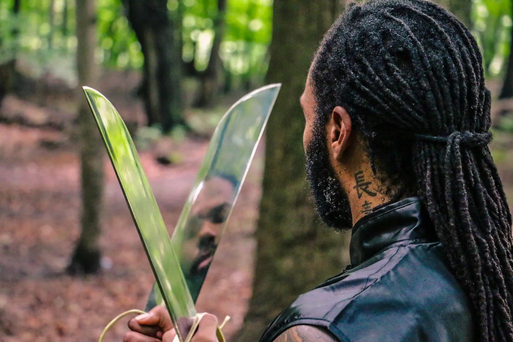 wing chun fighter holds two katanas in the woods. One of the katanas shows his reflection