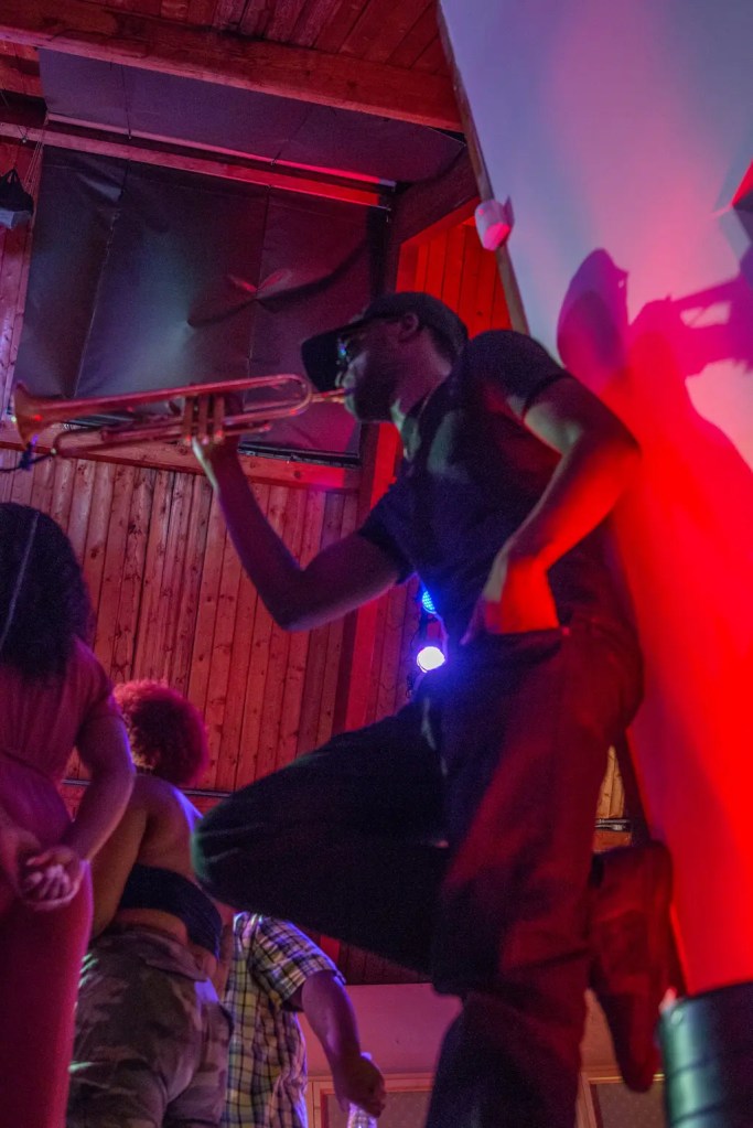 Rudy Ray leaning against the stage wall and playing the trumpet with his right hand, while his left hand rests in his jeans pocket