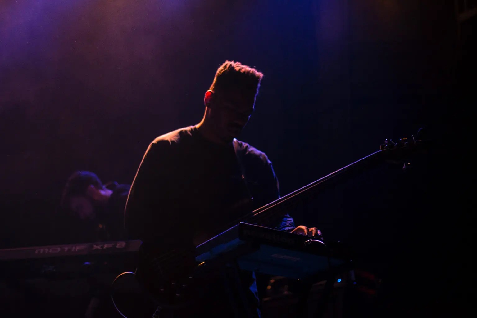 golden outline of a guitarist standing on stage holding his guitar with his right hand, and his left hand on a keyboard with a dark purple background