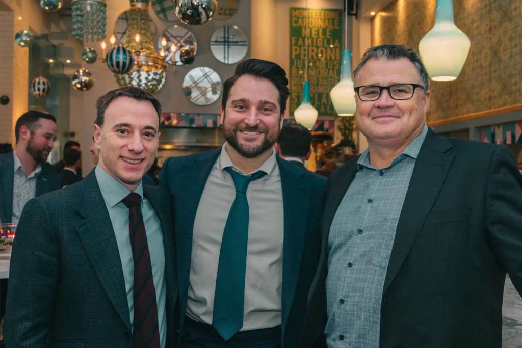 3 men in suits posing, at a festive setting with teal ornaments hanging in the background