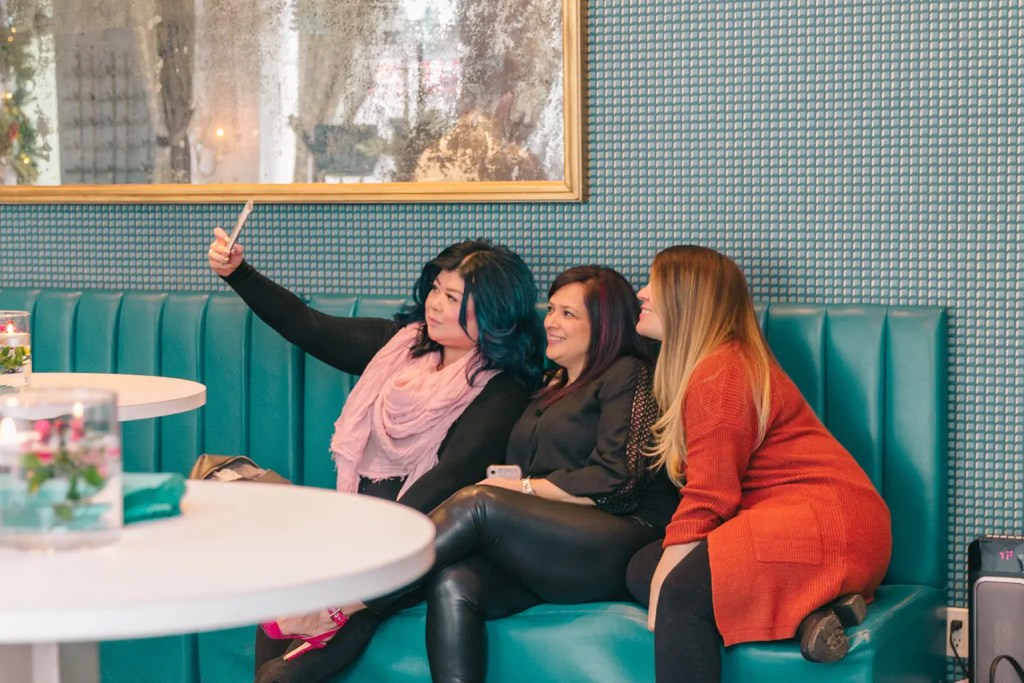 Three women huddled close together. The woman on the left is holding a mobile phone with her right hand and takes a selfie